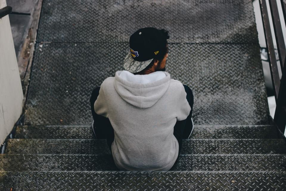 A man in a hooded jacket sits alone on outdoor steps, seen from behind.