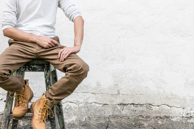 A man in work boots sits on a stool against a white wall.